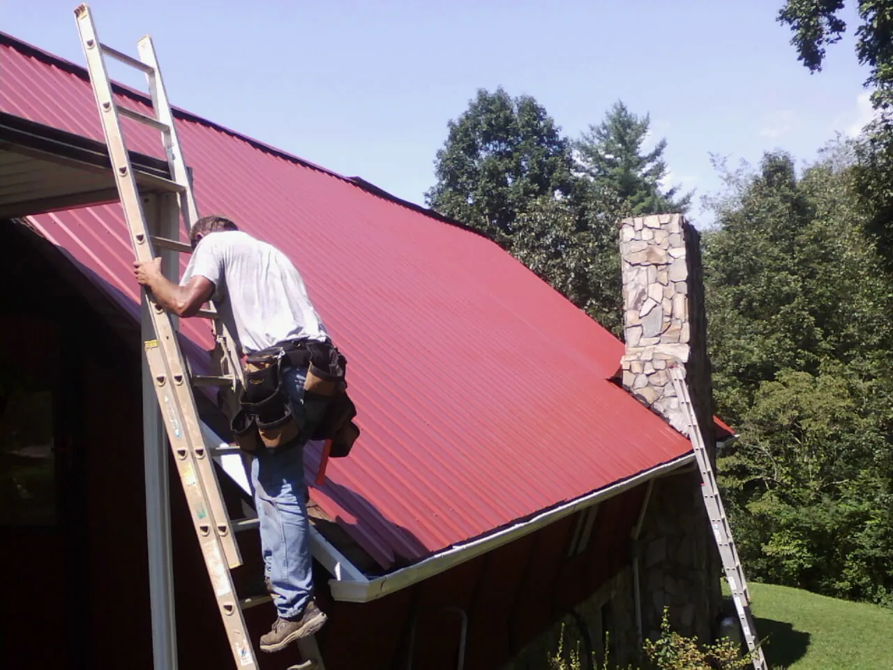 Roofer on a ladder beside a red metal roof and stone chimney — Soffit & Fascia Repair by Roofing Brothers in Mineral Ridge